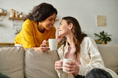İki kadın rahat bir kanepede içeceklerini yudumlarken hassas ve samimi bir anı paylaşıyor..