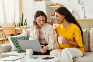 İki kadın evde keyifli bir anı paylaşıyor, içeceklerini yudumlarken bir dizüstü bilgisayarla bağ kuruyorlar..