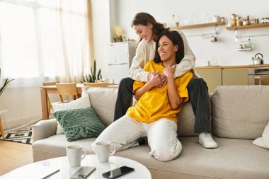 İki kadın kanepelerinde keyifli bir anı paylaşıyor, rahat ve samimi bir ev atmosferiyle çevrili..