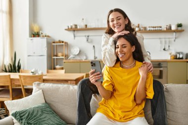 İki kadın evde neşeli bir anı paylaşıyor, kanepede sarılıyorlar ve birlikte telefona bakıyorlar..