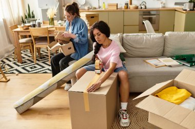 Kutularla ve gülümsemelerle çevrili iki kadın sevinçle yeni evlerini açıyor ve düzenliyor..