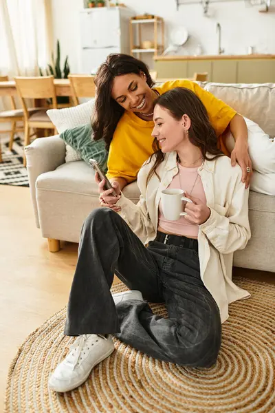 İki kadın rahat bir kanepede rahatlar, gülümsemelerini ve sıcak bir içkiyi paylaşırlar..