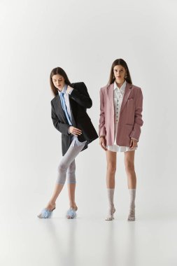 İkiz kız kardeşler, temiz bir arkaplan karşısında şık kıyafetler içinde poz verirken zarafet ve tarz sergiliyorlar..