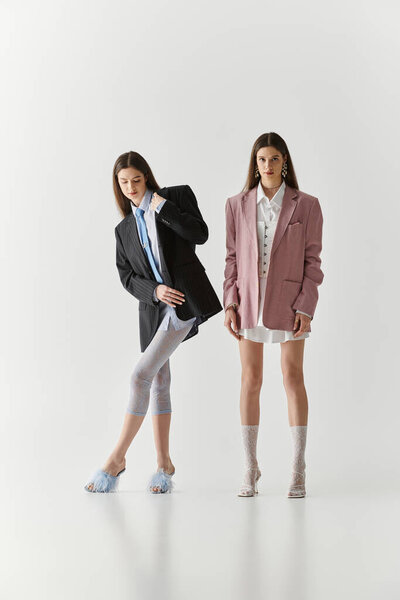 Сестры-близнецы излучают элегантность и стиль, позируя в модных нарядах на чистом фоне.