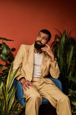 Sakallı genç adam yaz modasını sergiliyor. Canlı yeşillikler arasında, sıcak tonlarda oturuyor..