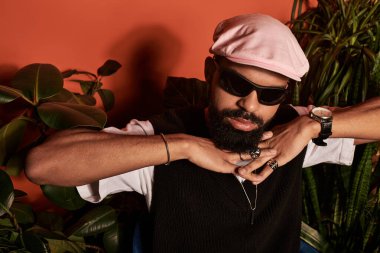 Genç bir adam yaz tarzıyla fiyakalı giyiniyor, yemyeşil bitkilerin arasında rahatlıyor..