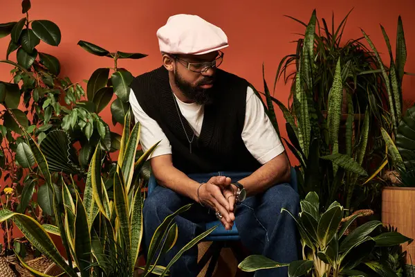 Moda kıyafeti giyen genç bir adam yemyeşil bitkilerin arasında oturur, rahat bir ortamda yaz titreşimleri yayar..