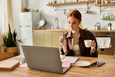 Kızıl saçlı kadın kahvesini yudumluyor ve mutfak masasında çalışıyor..