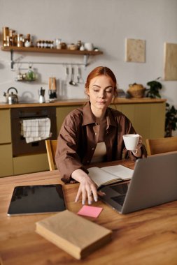 Kızıl saçlı kadın, parlak bir mutfakta kahvenin tadını çıkarırken dizüstü bilgisayarla meşgul oluyor..