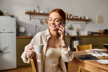 Güzel kızıl saçlı bir kadın sıcak bir içkiyi yudumlarken neşeli bir telefon görüşmesi yapmaktan hoşlanır..
