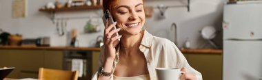 Gülümseyen kızıl saçlı kadın telefonda konuşuyor ve şık bir mutfakta kahvesini yudumluyor..