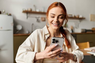 Kızıl saçlı kadın sıcak ve davetkar bir mutfak atmosferinde telefonuyla meşgul olurken gülümsüyor..