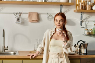 Kızıl saçlı neşeli bir kadın, parlak bir mutfakta dinlenirken sıcak bir içecek yudumluyor..