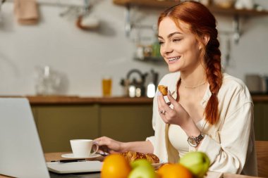 Kızıl saçlı bir kadın pastadan ve kahveden hoşlanır. Dizüstü bilgisayarına odaklanır..