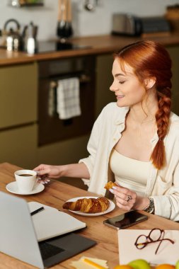 Kızıl saçlı bir kadın sıcak bir evde laptopuna odaklanırken kahve ve hamur işlerinin tadını çıkarıyor..