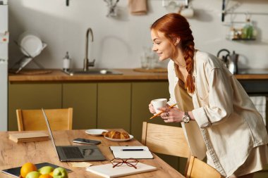 Parlak ışıklandırılmış mutfakta kızıl saçlı bir kadın kahvenin tadını çıkarıyor ve laptopuyla meşgul oluyor..