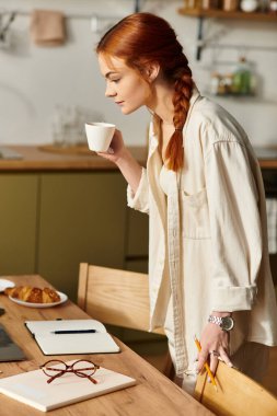 Güzel kızıl saçlı bir kadın kahvesini yudumluyor, parlak bir mutfak ortamında görevlerine odaklanıyor..