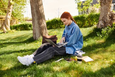 Yeşillik bir parkta, canlı kızıl saçlı bir kadın laptopuna yoğunlaşır ve etrafı doğa ile çevrilidir..