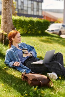 Canlı bir parkta dinlenen kızıl saçlı bir kadın, kahvesini yudumlarken dizüstü bilgisayarında çalışıyor..