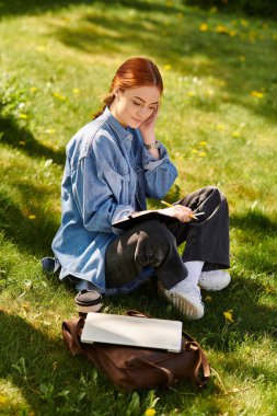 Kızıl saçlı kadın çimlerin üzerinde oturur, derinden güneşin altında defterine yazmaya odaklanır..