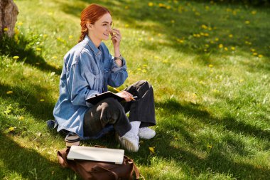 Neşeli kızıl saçlı bir kadın yeşil çimenlerde oturur, güneş ışığında güneşlenirken düşüncelerini yazar..