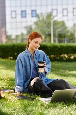 Kızıl saçlı kadın çimenlerin üzerinde kahvesini yudumluyor, rahat bir öğleden sonra laptopuna odaklanıyor..