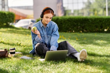 Kızıl saçlı bir kadın yumuşak çimenlerde oturur ve dışarıda dizüstü bilgisayarla eğlenir..