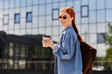 Parlak gülümseyen kızıl saçlı kadın şehirde elinde bir kahve fincanı tutarak güvenle yürüyor..