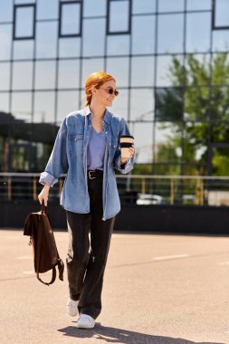 Kendine güvenen kızıl saçlı bir kadın, parlak güneş ışığı altında kahve ve sırt çantasıyla yürüyor..
