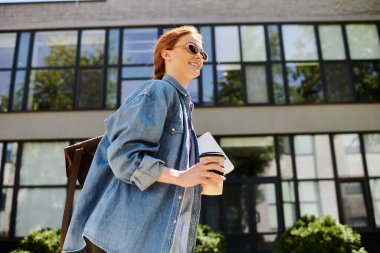 Neşeli kızıl saçlı bir kadın, işlek, güneşli bir sokakta, kendine güvenen bir şekilde kahveyle yürüyor..