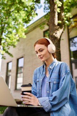 Kızıl saçlı, kulaklıklı bir kadın laptopuyla güneşli ağaçların altında çalışırken kahvesini yudumluyor..