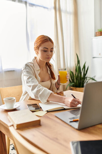 A woman with vibrant red hair writes notes while sipping juice, surrounded by a cozy kitchen.