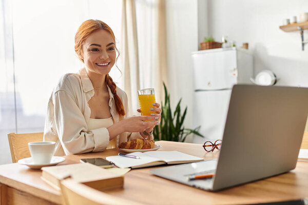 Cheerful red haired woman sips juice while working at a sunlit table in a cozy kitchen.