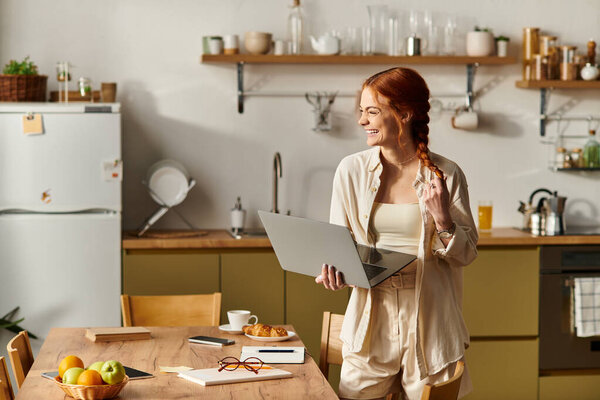 Brightly lit kitchen showcases a cheerful red haired woman engaged in creative work while smiling.