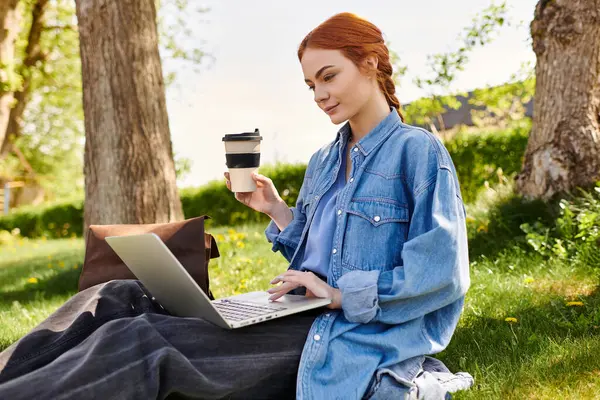 Kızıl saçlı kadın, sakin bir parkta dizüstü bilgisayarına odaklanırken çimenlerde oturup kahvesini yudumluyor..