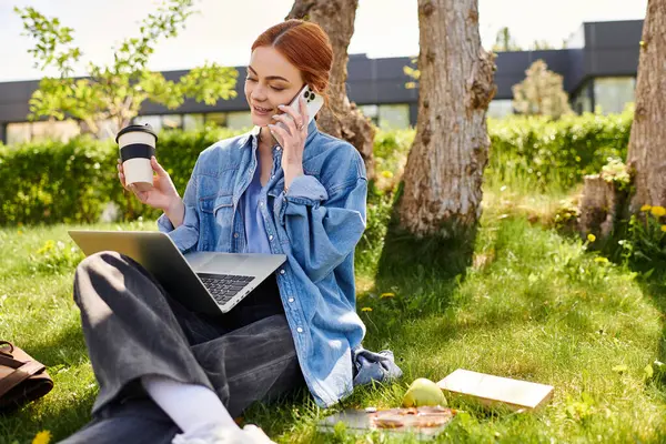 Kızıl saçlı bir kadın dışarıda laptopuyla çalışırken telefonundan kahve yudumluyor..