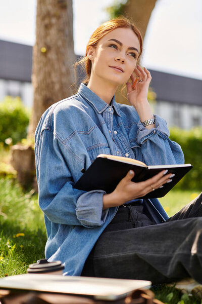 Красивая женщина с рыжими волосами созерцает и пишет в блокноте под солнцем в парке.