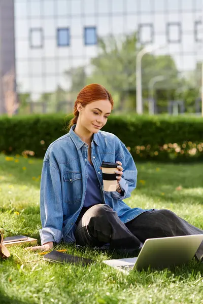 Kızıl saçlı kadın çimenlerin üzerinde kahvesini yudumluyor, rahat bir öğleden sonra laptopuna odaklanıyor..