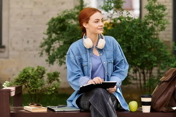 Parlak güneş ışığı, bankta oturan kızıl saçlı bir kadını aydınlatıyor, bir defterle ders çalışıyor..