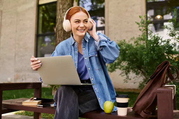 Kızıl saçlı kadın laptopunu kullanırken ve açık havada müzik dinlerken gülümsüyor..