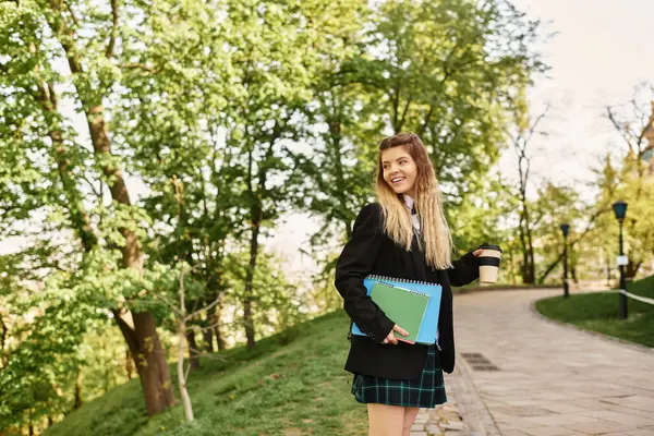 Güzel üniversiteli kız yeşil bir yolda yürüyor, gülümsüyor, elinde kahve ve defter tutuyor..