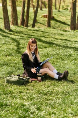 Genç yetişkinler yemyeşil çimlerde oturur, sakin bir parkta yanında defterlerle ders çalışmaya odaklanırlar..