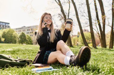 Neşeli bir üniversite öğrencisi çimenlerde oturur, kahvesini yudumlar ve telefonuyla sohbet eder, ders çalışır..