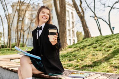 Üniversite öğrencisi kampüste kitap ve kahveyle rahatlıyor, büyüleyici üniformasıyla gülümsüyor..