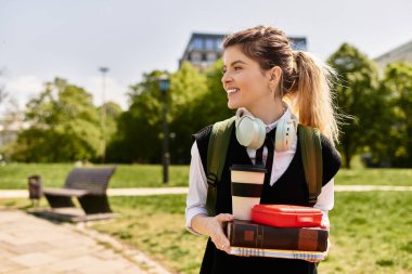 Genç yetişkin bir kız öğrenci elinde kitap ve kahveyle kampüste geziniyor..