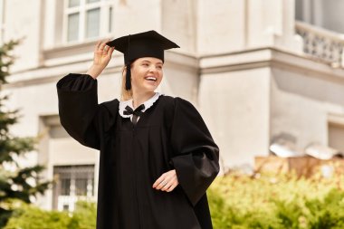 Mezuniyet elbisesi ve şapkası içinde genç bir kadın akademik başarısını dışarıda sevinçle kutluyor..