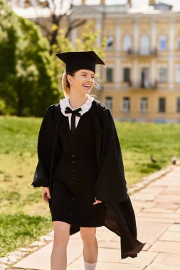Neşeli üniversiteli kız, mezuniyet elbisesi ve şapkasıyla kampüsteki yemyeşil alanda gururla yürüyor.