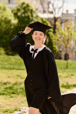 Genç bir yetişkin mezuniyetini güneşli bir ortamda gülümseyerek ve şapkasını sallayarak kutluyor..