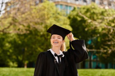 Bir üniversite mezunu ağaçların arasında elbisesi ve şapkasıyla sevinçle poz verir..