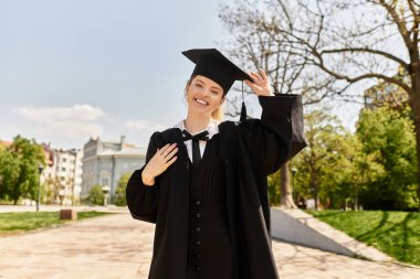 Parlak genç bir yetişkin, güzel bir parkta mezuniyet şapkasını düzeltirken ışıl ışıl gülümser..